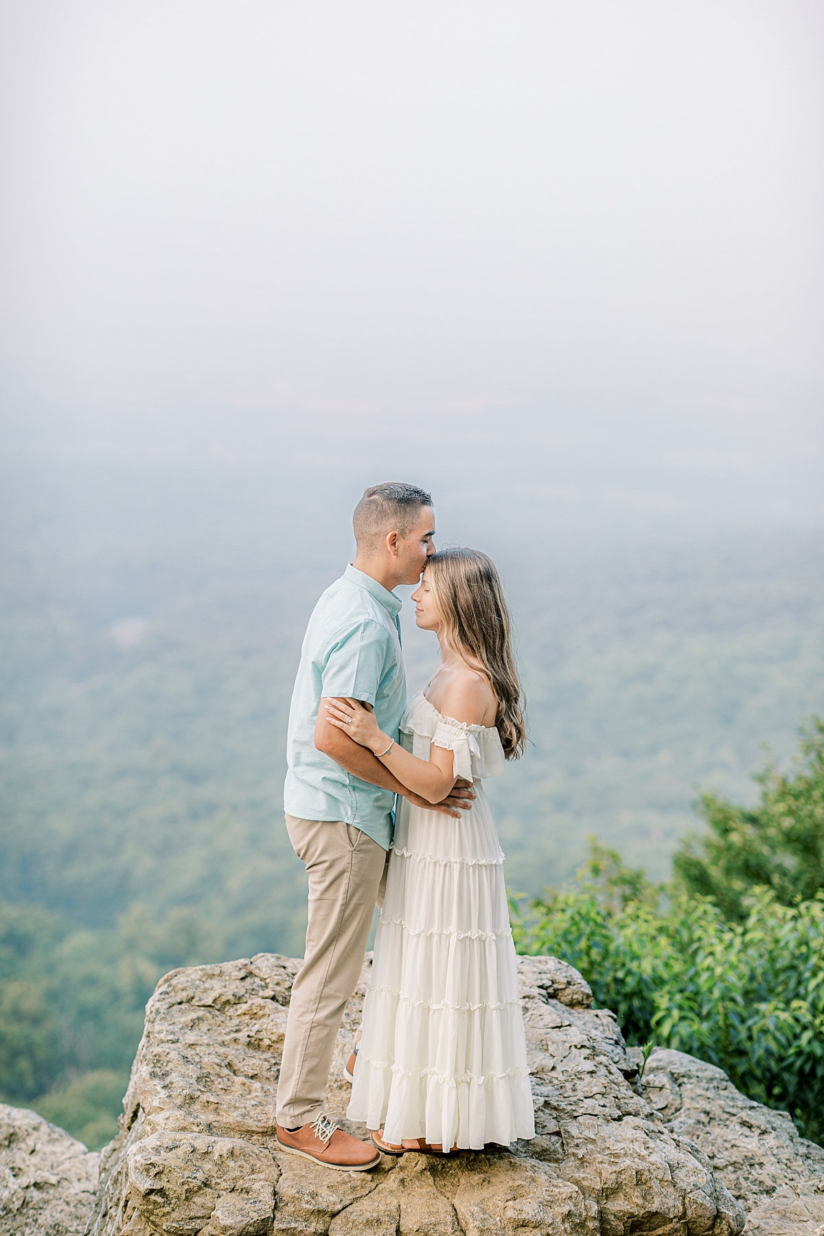 Engagement photo of a couple at Hawk Mountain Sanctuary in PA. The couple stands on rocks with a beautiful view as the groom kisses the bride's forehead